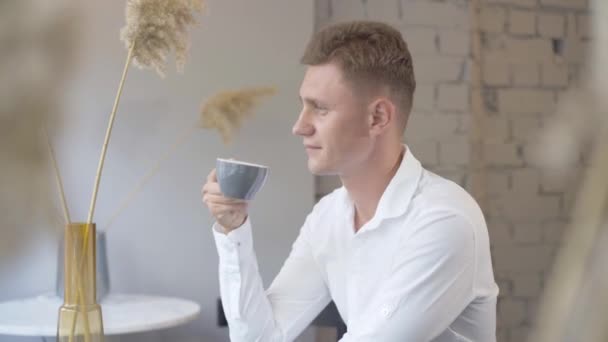 Joven empresario exitoso bebiendo café y mirando por la ventana en la cafetería. Hombre de negocios caucásico guapo seguro disfrutando del almuerzo en el interior y sonriendo. Estilo de vida empresarial. — Vídeos de Stock