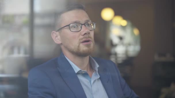 Primer plano de hombre de negocios caucásico confiado en gafas que hablan con una persona desconocida. Retrato de un joven guapo sentado en la cafetería detrás de una ventana de cristal y charlando. Estilo de vida empresarial. — Vídeos de Stock