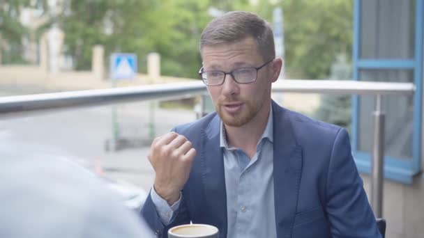 Pozitivní sebevědomý muž v brýlích a obleku sedí ve venkovní kavárně a mluví s nepoznatelným chlapem. Portrét úspěšného bělošského mladého podnikatele na obchodní schůzce. — Stock video