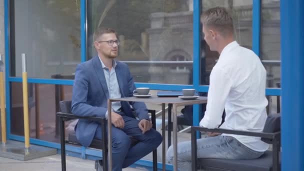 Deux hommes d'affaires caucasiens heureux serrant la main et parlant à la table dans un café extérieur. Des hommes confiants qui font affaire au déjeuner. Affaires et succès. — Video