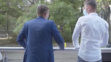 Erkek iş ortaklarının maskeli balkonda dikilip konuştuklarını gördüm. Covid-19 pandemik tecritte işbirliği hakkında kendinden emin genç beyaz adamlar konuşuyor..