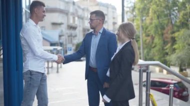 İş merkezinin yanında el sıkışan mutlu iş arkadaşları ya da ortakların yan görünüşü. Gözlüklü neşeli beyaz adam zafer jesti yapıyor. Başarılı ekibin portresi iş anlaşması yapıyor..