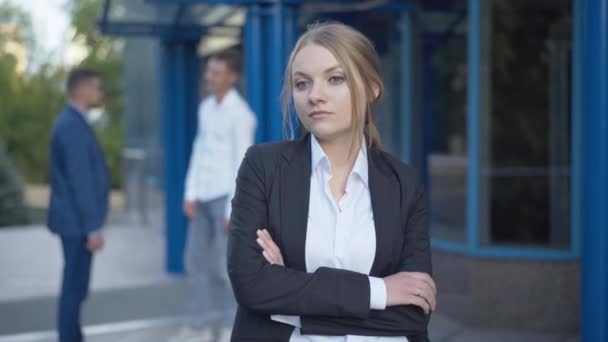 Retrato de una hermosa joven empresaria parada al aire libre con las manos cruzadas mientras dos hombres estrechan las manos al fondo. Mujer caucásica reflexiva esperando la reunión en la entrada del centro de negocios. — Vídeo de stock