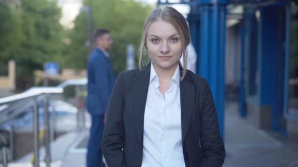 Hermosa joven empresaria posando en la entrada del centro de negocios con gente borrosa en el fondo. Retrato de mujer caucásica sonriente confiada de pie al aire libre y mirando a la cámara. — Vídeo de stock