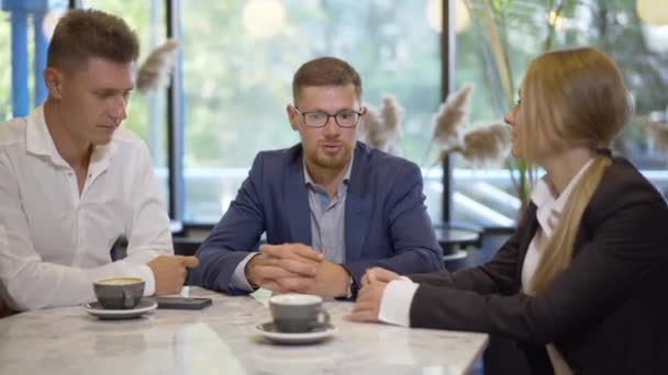 Drie succesvolle mensen zitten in café en praten. Portret van zelfverzekerde blanke mannen en vrouwen die praten over samenwerking bij koffiepauze. Teamwork van jonge zelfverzekerde ondernemers. — Stockvideo
