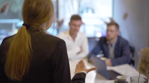 Vista posterior de la confiada empresaria entusiasta hablando con hombres borrosos sentados a la mesa en el fondo. Mujer caucásica joven irreconocible explicando plan a socios de negocios en la cafetería. — Vídeos de Stock