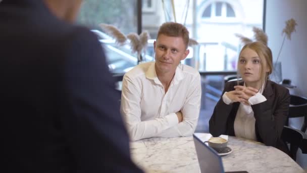 Ung vit man och kvinna i formella kostymer sitter på café och pratar med oigenkännlig kollega eller partner. Porträtt av självsäkra intelligenta affärsmän som träffas inomhus. — Stockvideo