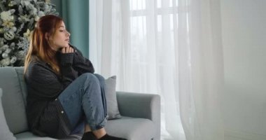 Yalnız genç bir kadın Noel 'i yalnız geçiriyor. Yeni yıl arifesinde kanepede oturup düşünen güzel kızıl saçlı beyaz kadın portresi. Yalnızlık kavramı. Sinema 4k ProRes Merkezi.
