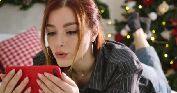 Πορτρέτο της απορροφημένη νεαρή γυναίκα κοιτάζοντας οθόνη smartphone και χαμογελώντας. Κοντινό πλάνο της απορροφημένης όμορφης κοκκινομάλλας που βλέπει ταινία στο διαδίκτυο τα Χριστούγεννα. Κινηματογράφος 4k ProRes HQ. — Αρχείο Βίντεο