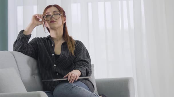 疲れ切った若いフリーランスの肖像画が眼鏡と思考を離陸。若い疲れた美しい女性が自宅でノートパソコンとソファに座っている。リモートワークの概念. — ストック動画