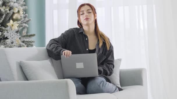 Перегруженная работой женщина закрывает ноутбук и растягивается, сидя на диване в канун Нового года. Портрет измученной юной кавказки, работающей в интернете на Рождество. Переработанная концепция. — стоковое видео