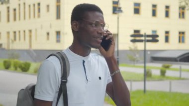 Arka planda gezinen küçük bir kız olarak telefonda konuşan Afro-Amerikan üniversite öğrencisinin yakın çekimi. Gözlüklü, sırt çantalı, dışarıda duran mutlu genç adamın portresi..