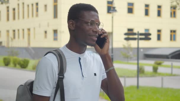 Close-up van een positieve Afro-Amerikaanse student die aan de telefoon praat als een klein meisje dat naar de achtergrond wandelt. Portret van een gelukkige jongeman in een bril met rugzak buiten. — Stockvideo