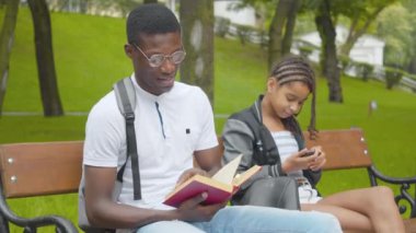 Güneşli parkta kitap okuyan Afrikalı Amerikalı bir adam arka planda selfie çeken küçük bir kız gibi. Neşeli erkek kardeş akıllı telefona gülümsüyor. Nesiller konsepti.
