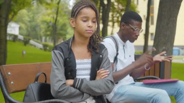 Porträt eines süßen beleidigten Mädchens mit Afro-Zöpfen, das mit verschränkten Händen sitzt, während der ältere Bruder im Hintergrund redet. Konflikt afroamerikanischer Geschwister im sonnigen Park. — Stockvideo