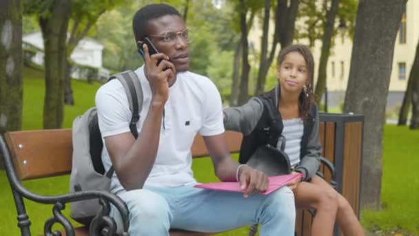 Busy African American man in eyeglasses talking on the phone ignoring pretty little girl. Portrait of serious brother and bored sister sitting on bench in sunny park outdoors. Family conflict. — Stock Video