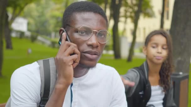 Nahaufnahme eines afroamerikanischen Mannes, der als gelangweiltes kleines Mädchen am Telefon versucht, seine Aufmerksamkeit auf sich zu ziehen. Umtriebiger Bruder ignoriert Schwester im sonnigen Park draußen. Konflikt zwischen Geschäftigkeit und Freizeit. — Stockvideo