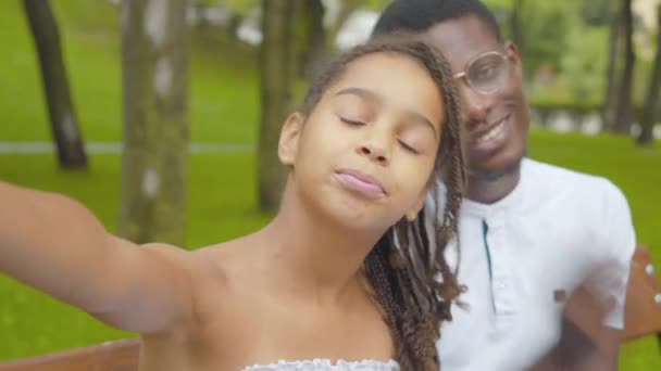 Point de vue selfie, jolie fille afro-américaine gaie prenant des photos avec un homme flou en arrière-plan. Sœur souriante s'amuser avec son frère dans un parc ensoleillé en plein air. — Video