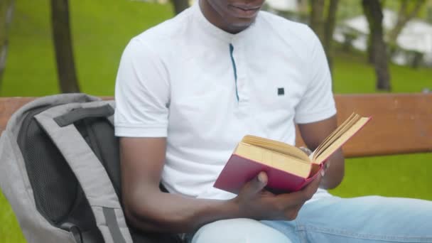 Nerozpoznatelný africký student vysoké školy čte nahlas ve slunečném parku. Soustředěný mladý muž sedící na lavičce s knihou a batohem. Životní styl a hobby koncept. — Stock video
