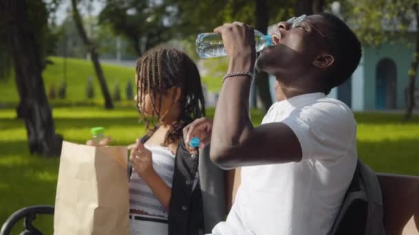Seitenansicht eines unbeschwerten afroamerikanischen Mannes, der im sonnigen Park erfrischendes Wasser trinkt und im Hintergrund Teenager-Mädchen, die Saft genießen. Entspanntes Geschwisterpaar ruht sich an Sommertagen im Freien aus. — Stockvideo