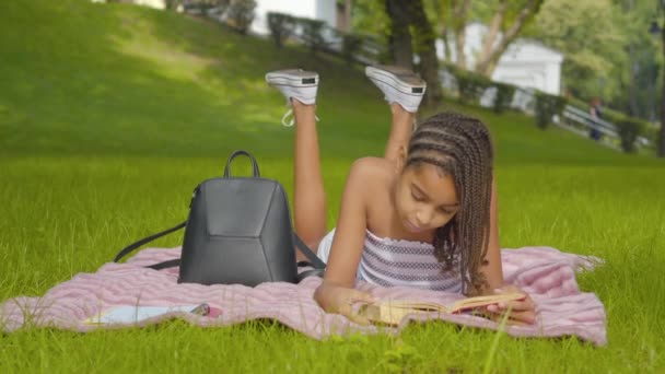 Parktaki yeşil çayırda uzanmış kitap okuyan konsantre bir kızın geniş açılı görüntüsü. Parkta güneşli bir günün tadını çıkaran Afro-Amerikan öğrenci portresi. Hobi konsepti. — Stok video