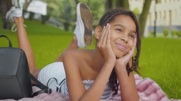 Portret van een mooi gelukkig meisje liggend op een zonnig weitje en lachend. Positieve Afro-Amerikaanse kind rust buiten op zomerdag. Jeugd- en geluksconcept. — Stockvideo