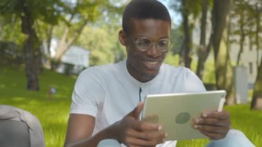 Telsiz iletişimi için tablet kullanan neşeli Afro-Amerikan bir adamın orta kare portresi. Mutlu genç adam konuşuyor ve dışarıda güneşli bir parkta oturmuş selfie kamerasına işaret ediyor. Modern yaşam tarzı.