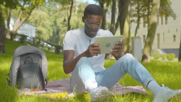 Portrét Afroameričana, jak odkládá tabulku, sundává si brýle a mnul si oči. Široký záběr mladého muže unaveného internetem a zařízeními sedícími ve slunečném parku. Koncept moderního životního stylu. — Stock video