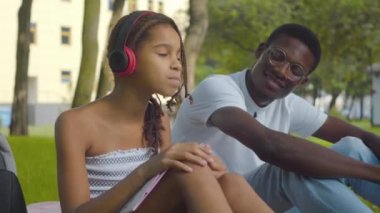 Neşeli şirin kızın portresi kulaklıkla müzik dinleyen ve gülümseyen Afrikalı Amerikalı genç adama kulaklık veren kız. Olumlu ablalar hafta sonları güneşli yaz parkında dinleniyorlar..