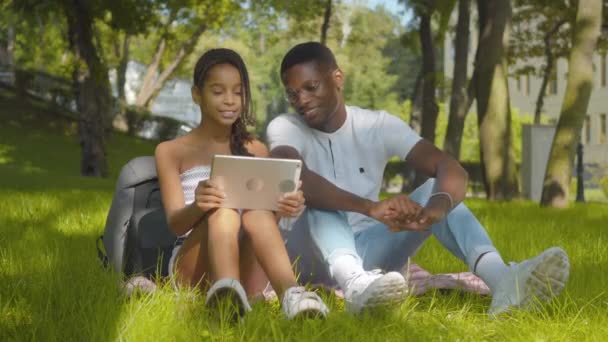 Sonriente joven afroamericano y niña usando tableta al aire libre y hablando. Retrato de hermano positivo y hermana navegando por Internet o viendo películas en línea en el soleado parque de verano. — Vídeo de stock