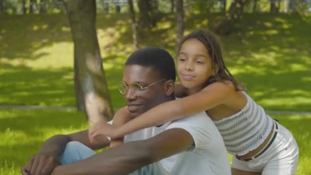 在阳光灿烂的夏季公园里,一个可爱的女孩拥抱男人的侧视图.描绘了快乐的非洲裔美国人姐姐和哥哥看着相机和微笑的形象。快乐与休闲的概念. — 图库视频影像