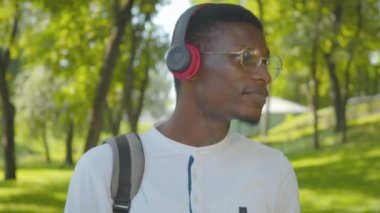 Gözlüklü, kulaklıklarını çıkarıp kameraya gülümseyen mutlu genç adamın yakın çekimi. Yazın güneşli bir parkta eğlenen neşeli Afro-Amerikalı adamın portresi. Boş zaman ve hobi konsepti.