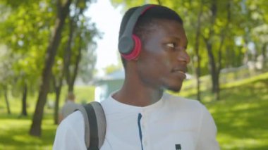 Kulaklıkla müzik dinleyen, kulaklıklarını çıkaran ve kameraya gülümseyen yakışıklı Afro-Amerikan bir adamın yakın plan portresi. Mutlu adam yaz parkında güneşli günün tadını çıkarıyor..