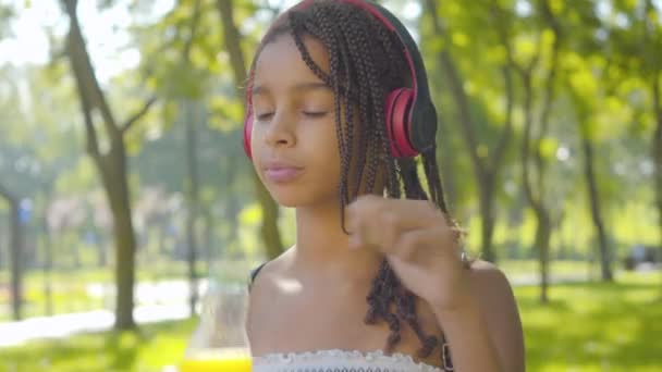 Nahaufnahme eines positiven kleinen Mädchens mit Kopfhörern, das im sonnigen Park Orangensaft trinkt. Porträt eines entspannten, fröhlichen afroamerikanischen Kindes, das sich an einem Sommertag im Freien ausruht. Freizeit- und Erholungskonzept. — Stockvideo