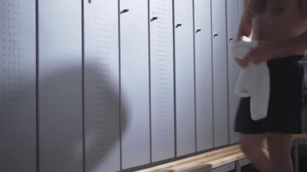 Young handsome Caucasian sportsman with naked torso entering dressing room, sitting down and rubbing face with white towel. Portrait of exhausted man leaning against locker in gym after workout. — Stock Video