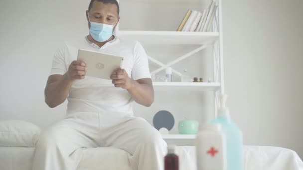 Porträt eines kranken Afroamerikaners mit Gesichtsmaske, der auf einer Krankenstation sitzt und Tabletten nimmt. Schwacher entspannter Patient spielt Videospiele in medizinischer Klinik zur Covid-19-Pandemie-Quarantäne. — Stockvideo