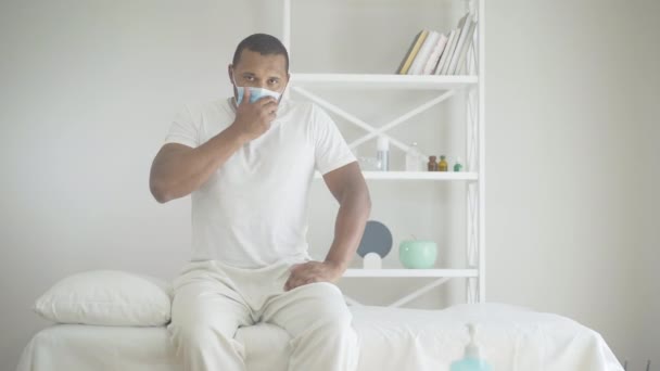 얼굴에 마스크를 쓴 아프리카계 미국인 청년이 병실 침대에 앉아 기침을 하고 있습니다. 코비 디온 -19 호흡기 질환을 앓고 있는 환자의 모습. 코로나 바이러스 대유행 개념. — 비디오