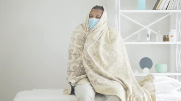 Un hombre triste envuelto en una manta en la sala del hospital. Retrato de un paciente afroamericano enfermo con fiebre Covid-19. Síntomas de la enfermedad por coronavirus. — Vídeo de stock