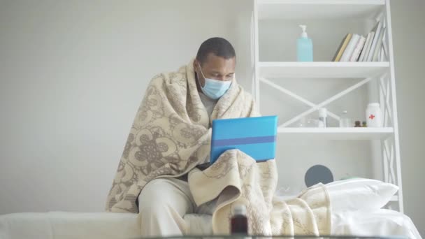 Ένας άντρας τυλιγμένος σε μια κουβέρτα που κλείνει το λάπτοπ και βγάζει τη μάσκα προσώπου. Πορτρέτο εξαντλημένων αφροαμερικανών ασθενών που εργάζονται στο διαδίκτυο από το νοσοκομείο για την πανδημία Covid-19. Τρόπος ζωής του Coronavirus. — Αρχείο Βίντεο