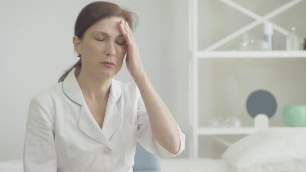 Porträtt av stressad läkare med huvudvärk hålla tempel med händer och suckar. Trött kaukasisk kvinna med symtom på Covid-19 infektionssjukdom efter arbete med sjuka patienter. — Stockvideo