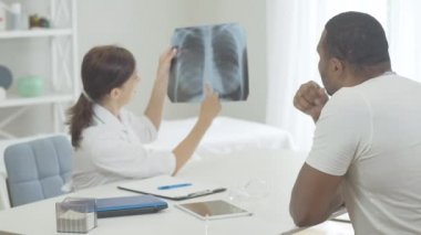 Afrika kökenli Amerikalı hastaya akciğer röntgenindeki komplikasyonları açıklayan profesyonel bir beyaz doktor. Hastanede hasta adamla konuşan kendine güvenen bir kadın. Kronik hastalık veya sigara içmenin sonucu.