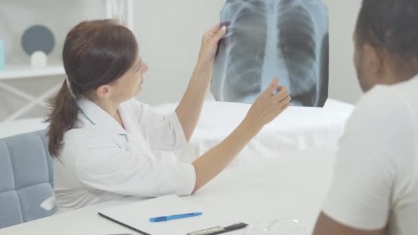 Medicul și pacientul examinează radiografiile pulmonare în spital. Bărbat afro-american și femeie caucaziană stând la masă și vorbind. Medicul consultă un tip bolnav cu boli respiratorii sau cancer . — Videoclip de stoc