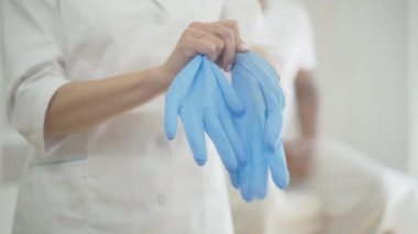 Doktor arka planda bulanık bir adam otururken koruyucu eldiven takıyor. Covid-19 küresel salgın sırasında tıbbi muayenede kişisel koruma kullanan tanınmayan bir kadın..