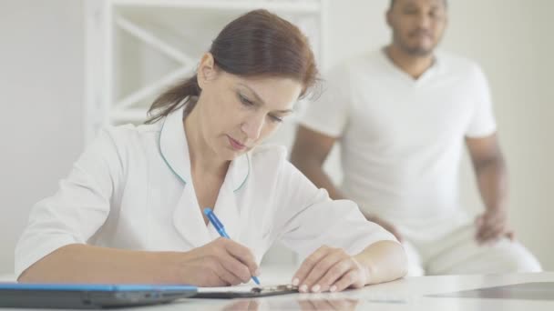 Médico serio concentrado llenando la historia clínica con el hombre preocupado borroso en el sofá de examen en el fondo. Retrato de mujer caucásica segura que trata al hombre afroamericano en el hospital. — Vídeos de Stock