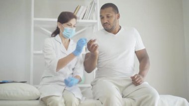 Kadın doktor yüksek ateşi olan hastadan termometre alıyor. Yüz maskesi takmış beyaz kadın portresi ve koruyucu eldivenler. Covid-19 salgınında Afrikalı Amerikalı bir adama danışıyor..