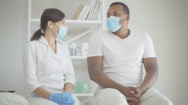 Yüz maskeli doktor ve hasta hastane koğuşunda oturmuş konuşuyorlar. Covid-19 'da Afrikalı Amerikalı bir adamı sakinleştiren destekleyici beyaz kadın portresi.