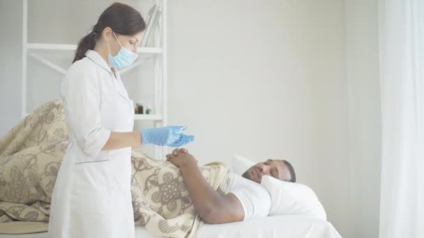 병원에서 남성 아프리카계 미국인 환자의 체온을 측정하는 여성 캅카스 의사. 얼굴 마스크를 하고 보호 장갑을 끼고 있는 전문직 여성이 온도계를 보면서 침대에 누워 있는 남자처럼 말하는 모습. — 비디오