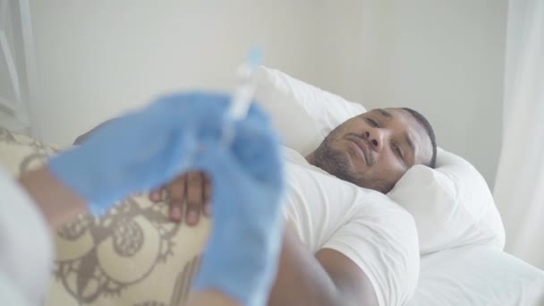 Hombre afroamericano enfermo agotado acostado en la cama y mirando a una mujer irreconocible con guantes protectores preparando la inyección en jeringa. Paciente tratado por enfermedad en la sala de hospital. — Vídeo de stock