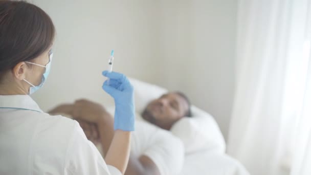 Professioneller Arzt oder Krankenpfleger mit Mundschutz und Schutzhandschuhen bereitet Spritze für die Injektion vor, während im Hintergrund verschwommener Patient auf dem Bett liegt. Kaukasische Frau behandelt afroamerikanischen Mann. — Stockvideo