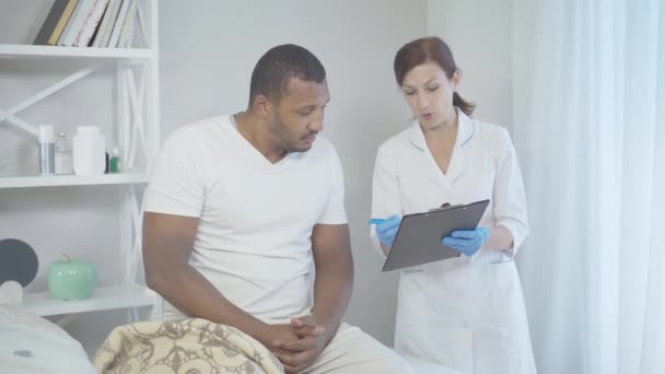 Dorosła biała kobieta rozmawiająca z mężczyzną siedzącym na łóżku na oddziale szpitalnym i uśmiechnięta. Portret profesjonalnego lekarza konsultującego się z afroamerykańskim pacjentem. Koncepcja zawodu lekarza. — Wideo stockowe